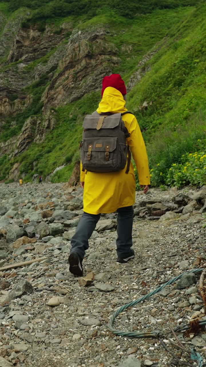 Persona con impermeabile giallo e zaino che cammina su una costa rocciosa.