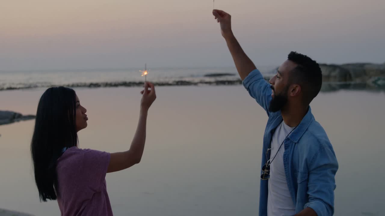 una joven pareja de raza mixta divirtiéndose con chispas en la playa 4k