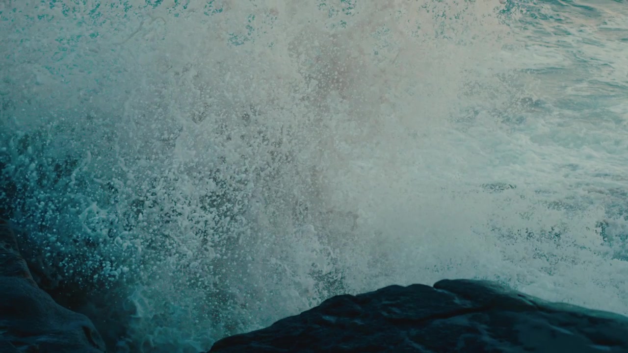 Explosive slow-motion shot of a powerful wave bursting against jagged dark rocks, spray erupting into deep teal water, raw coastal energy, dramatic ocean seascape.
