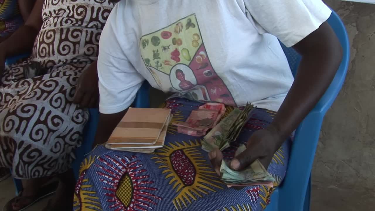Women in a Ghanaian village savings and loan group sit together counting cash and holding passbooks during a microcredit meeting