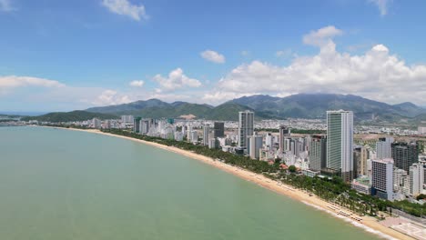 Große-Luftdrohne-Von-Nha-Trang-An-Einem-Sonnigen-Sommertag-Mit-Der-Kamera,-Die-Sich-In-Richtung-Der-Küste-Und-Des-Weißen-Sandstrandes-Bewegt,-Während-Der-Verkehr-An-Den-Hohen-Wolkenkratzer-Wohngebäuden-Vorbeifährt