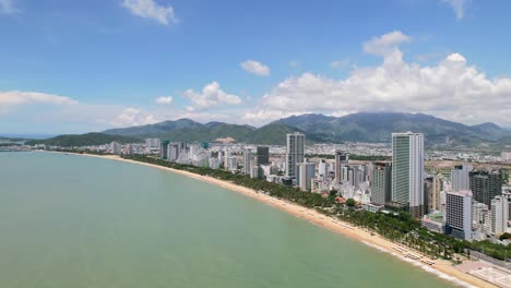 un amplio dron aéreo de nha trang en un soleado día de verano con una cámara moviéndose hacia la costa y la playa de arena blanca mientras el tráfico pasa por los altos rascacielos de los edificios residenciales