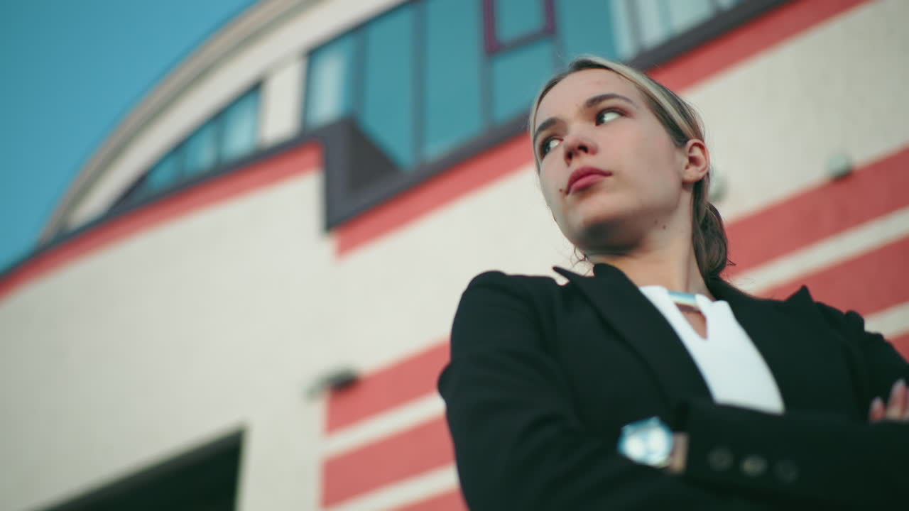 Young damsel in black blazer stands with arms crossed, gazing into distant sky with confident expression in front of modern curved building