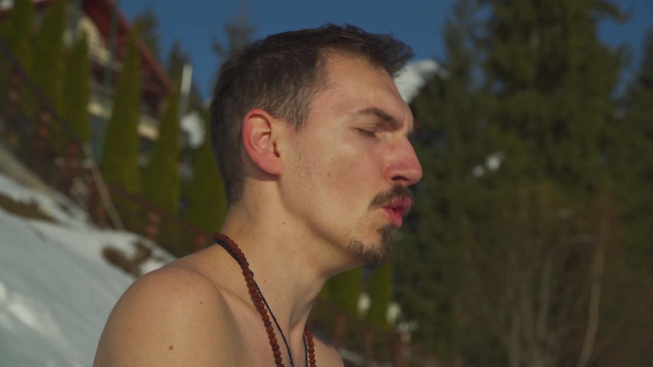 눈 덮힌 산 풍경에 깊이 앉아 숨을 들이마시고 내쉬는 셔츠 없는 남자의 얼굴을 가까이서 볼 수 있습니다.