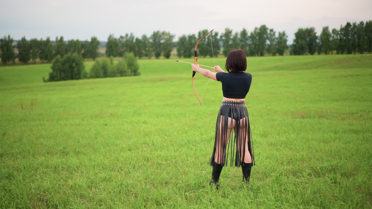 woman archer in green meadow standing back to camera drawing bow, leather fringe skirt and boots, slow deliberate stance, aim and release motion, overcast sky and distant tree line, calm solitary