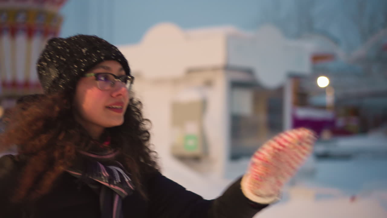 Female skater in winter hat and glasses enjoys outdoor skating during snowy evening, smiling while dressed in warm jacket and scarf with amusement park lights and snowy background creating festive atmosphere