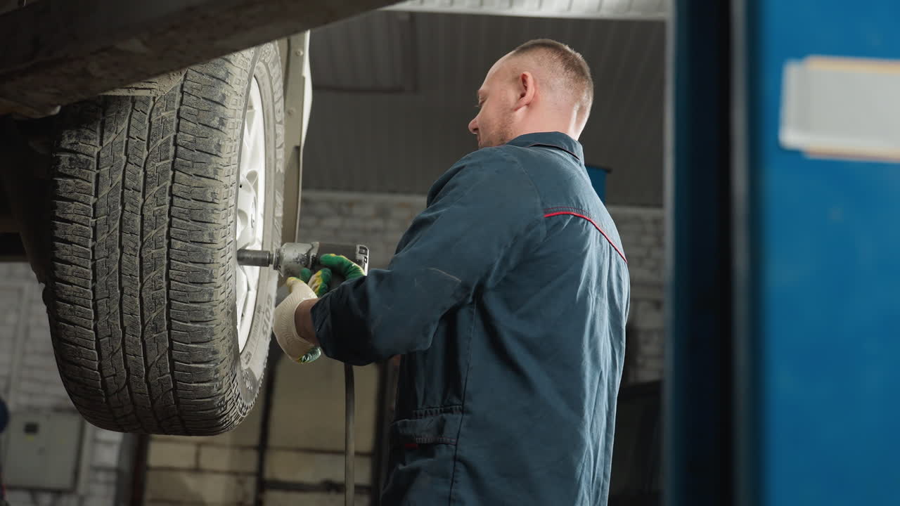 vista aérea de un mecánico en uniforme azul usando una herramienta neumática para apretar los pernos de las ruedas de un automóvil levantado en el taller, mientras que el compañero se acerca, observa el trabajo y asume en aprobación