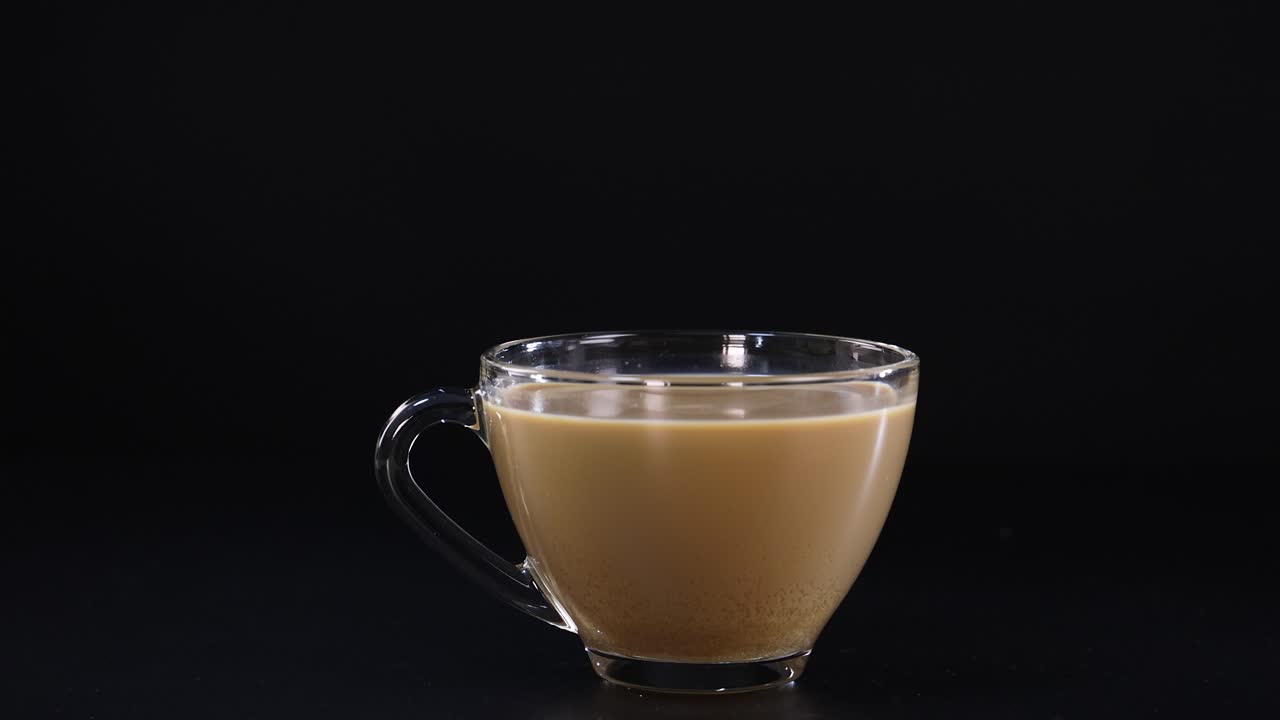 A hand pours sugar into a cup of coffee against a black background, highlighting the granules in motion