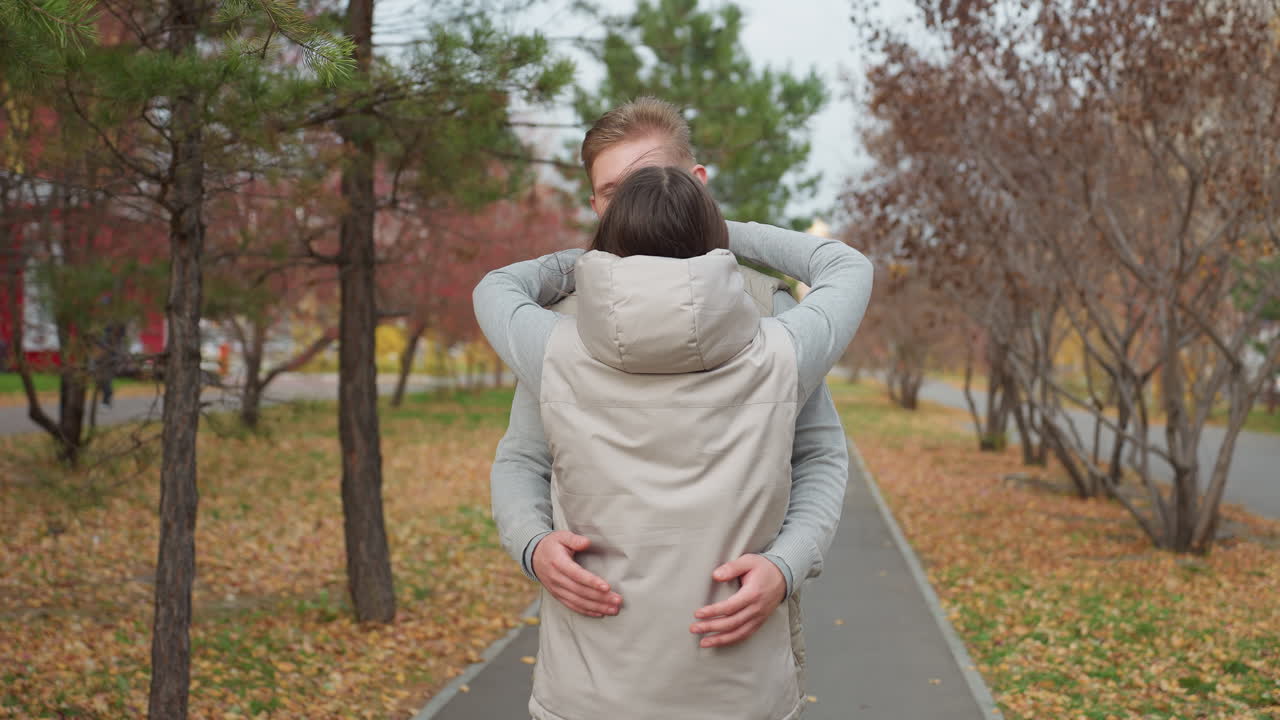 Back view of woman approaching her brother on park path lined with autumn trees as they prepare for warm embrace with leaves scattered on ground and gentle breeze moving through foliage