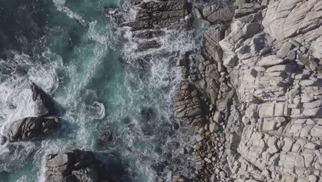 Descending-rotating-birdseye-aerial-view-above-rocky-beach-splashing-rough-blue-sea-waves-shoreline