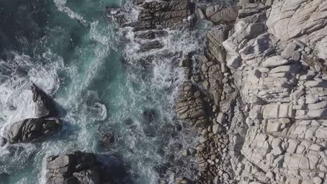 descending rotating birdseye aerial view above rocky beach splashing rough blue sea waves shoreline