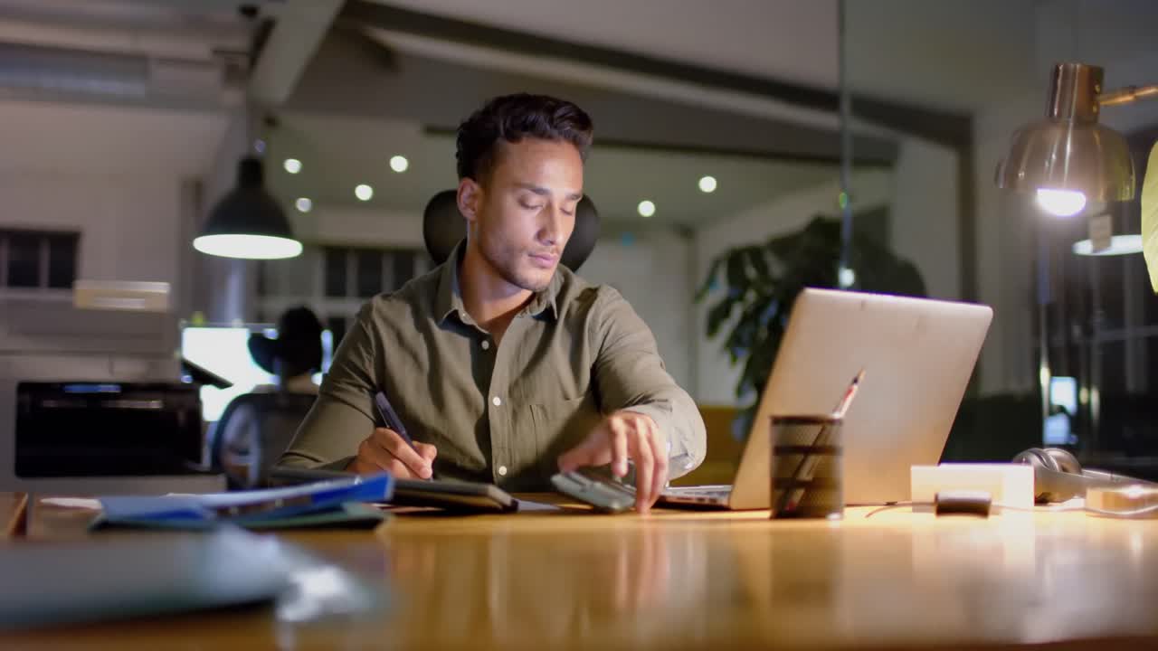 Biracial businessman at desk using laptop and smartphone in office at night, slow motion