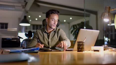 Biracial-businessman-at-desk-using-laptop-and-smartphone-in-office-at-night,-slow-motion