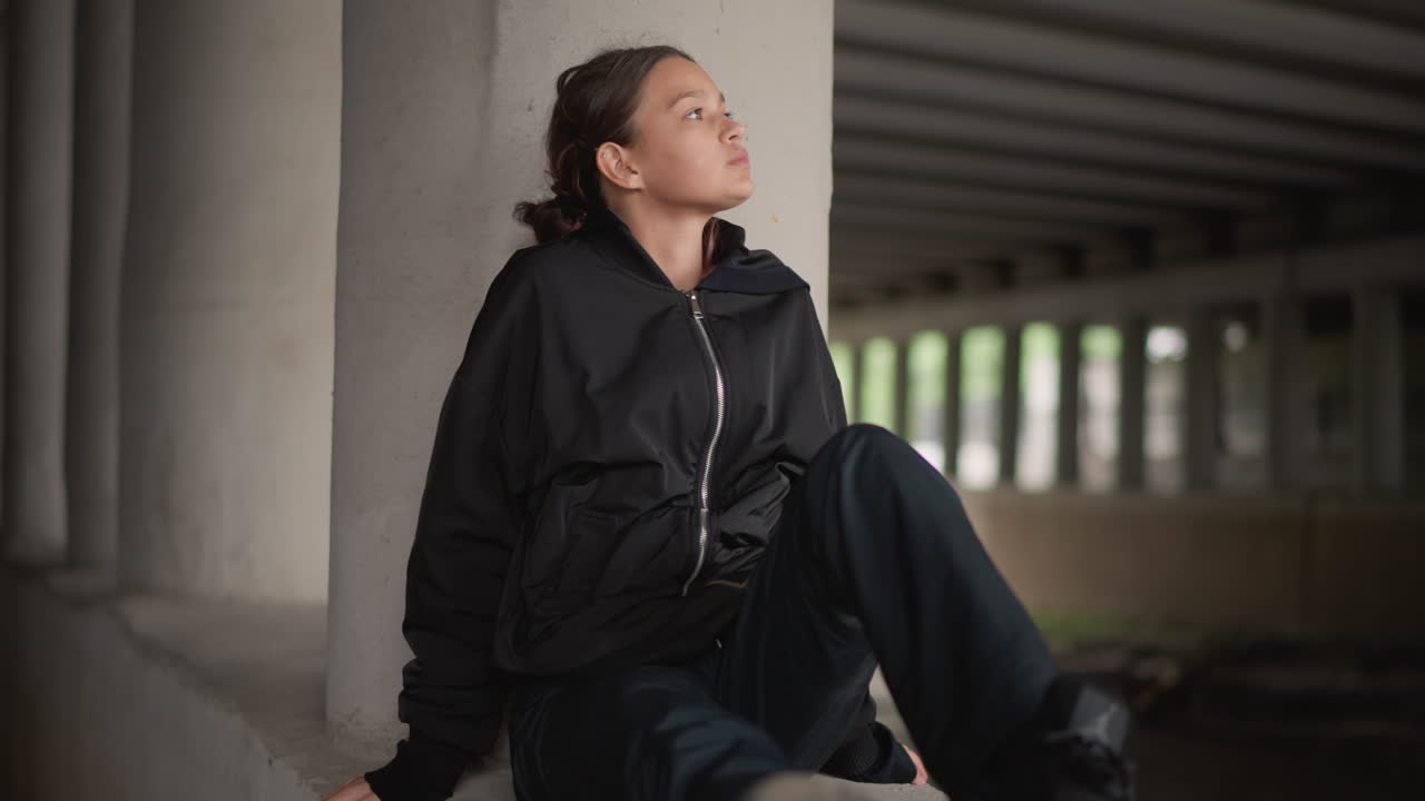 Young woman sitting on concrete ledge, commuter holding jacket and scanning street from rainsheltered underpass, boots on damp surface, candid cinematic portrait with soft bokeh and muted city tones