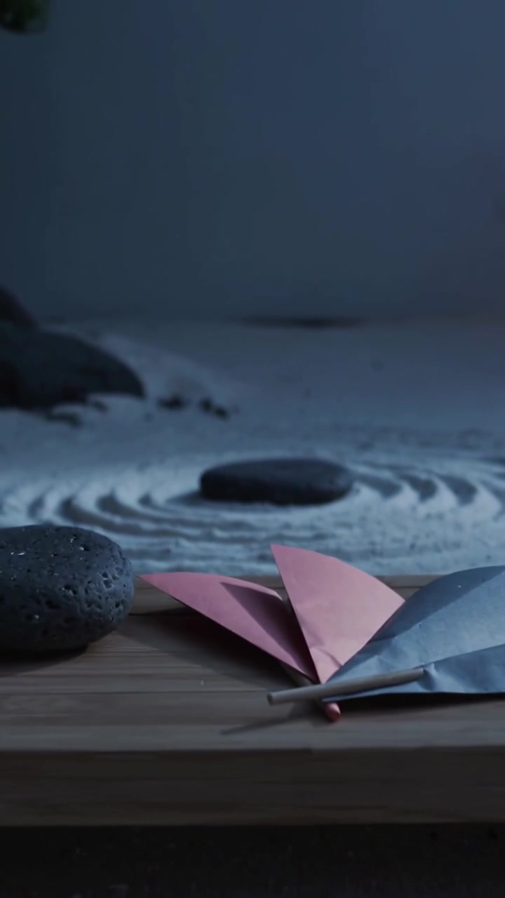 Vertical video: Pulling camera back revealing dark stone on raked sand, showing tray and origami