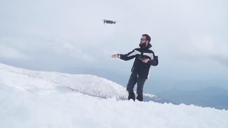 Un-Hombre-Con-Barba-Lanza-Un-Dron-Desde-La-Mano-Mientras-Está-Parado-En-La-Nieve-En-Lo-Alto-De-La-Montaña