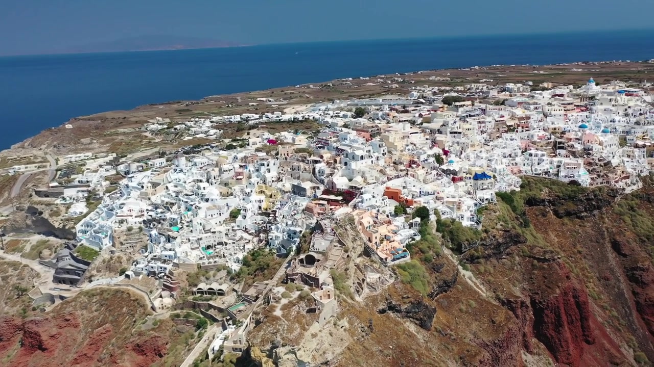 Bela tomada ampla de Oia, Santorini, apresentando casas brancas icônicas, igrejas de cúpulas azuis e dramáticas falésias vulcânicas vermelhas que descem em direção ao azul profundo do Mar Mediterrâneo em um dia claro.