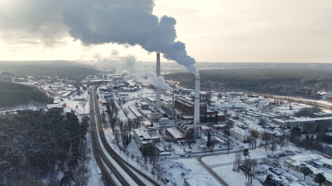 Drone view of Vilnius combined heat and power plant in snowy winter, tall chimney releasing steam plume over forest and city outskirts