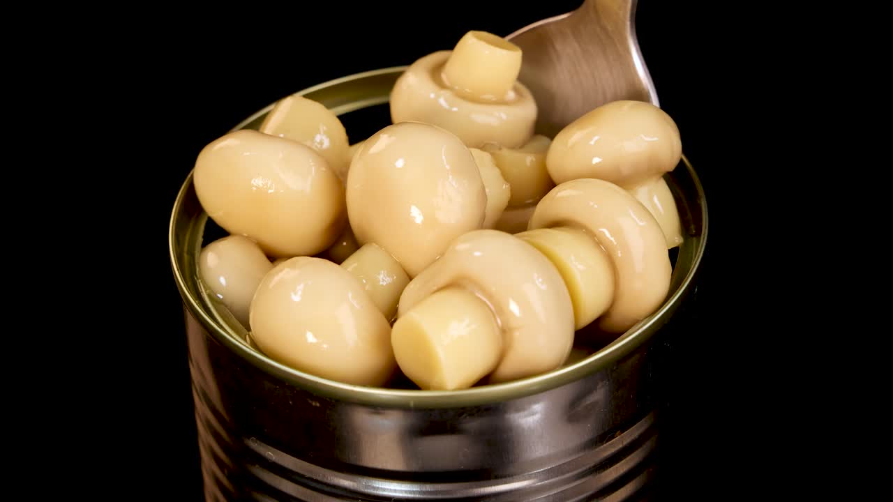 A metal spoon lifts whole champignon mushrooms from an open can against a black background, with even studio lighting and close-up perspective