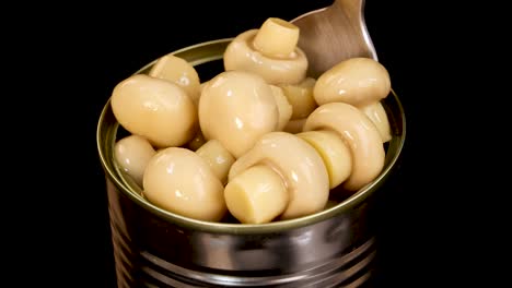 a metal spoon lifts whole champignon mushrooms from an open can against a black background, with even studio lighting and close-up perspective
