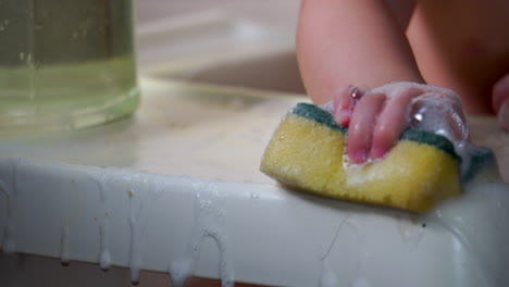 Lindo-Niño-Sentado-En-Una-Trona-Limpiando-Con-Una-Esponja-Jabonosa-Y-Agua-En-La-Mesa-Blanca