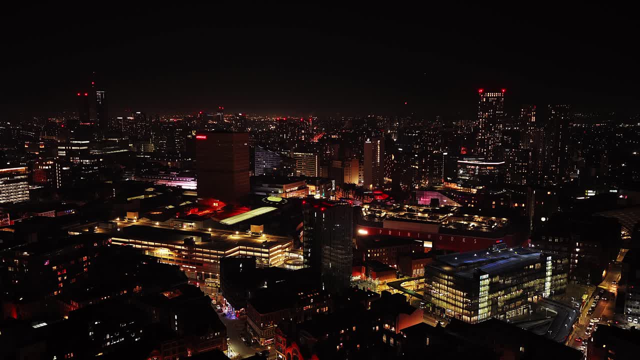 Nighttime aerial image shows Manchester’s city centre with Beetham Tower illuminated, dense traffic on Deansgate and Oxford Street, and surrounding high-rise buildings glowing under urban lighting