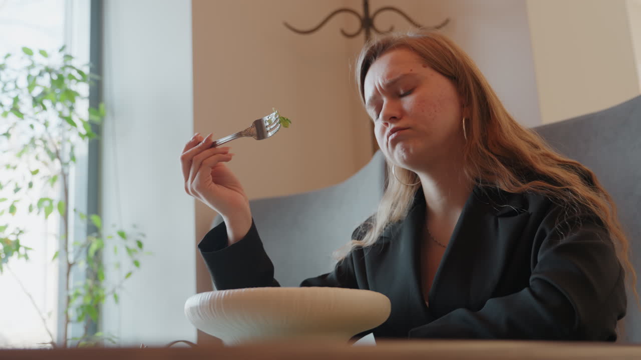 Kaukasisk kvinde tøver over salat ved vinduet, studerende studerer gaffelfuld mod plantebaggrund, blødt dagslys, eftertænksomt udtryk, diskret hovedvip, moderne caféinteriør, beslutningsøjeblik