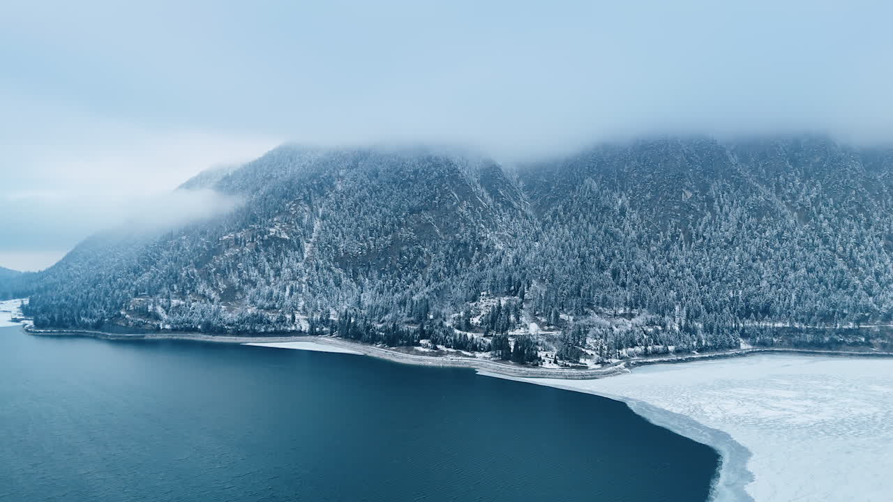 Tops of the steep mountains covered with woods are hiding in the grey cloudscape. Lake at the rock foot is partially in ice. Aerial view.