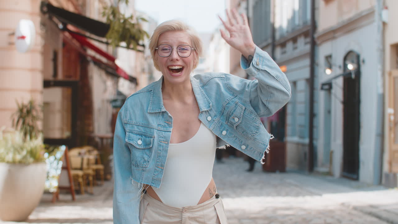 Cheerful young blonde woman smiling friendly at camera waving hands gesturing on city urban street