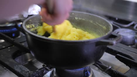 closeup of female cook stirring corn meal with wooden spoon in black pot on gas stove, woman preparing traditional polenta in kitchen