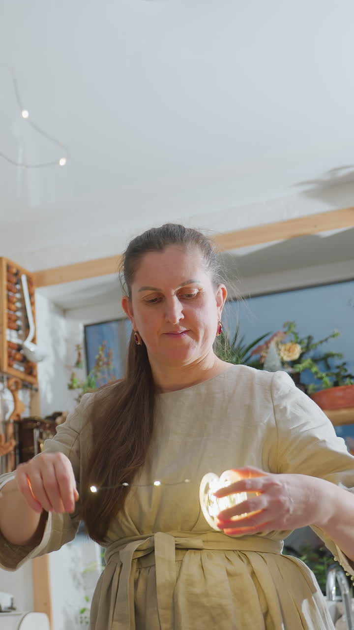 mujer caucásica colgando luces de ambiente cálidas en una cocina soleada, pasando luces a través de una estantería de madera, ajustando una esfera brillante, comprobando una bombilla y sonriendo, llevando un delantal de lino y un vestido casual