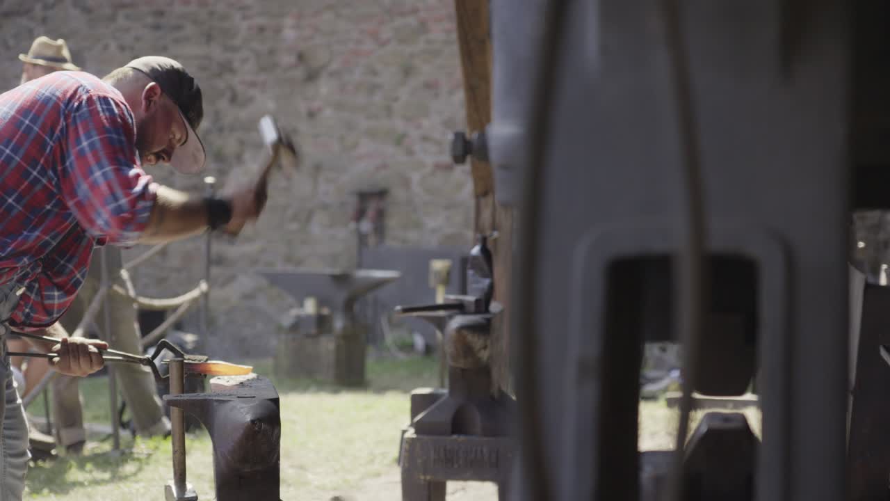 A blacksmith hammers hot iron on an anvil in an outdoor workshop.