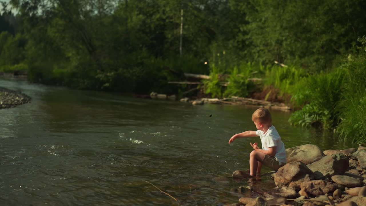 Little boy entertains on riverside throwing stone into lake during summer season. Cute toddler child spends time enjoying fantastic view of reservoir on sunny day