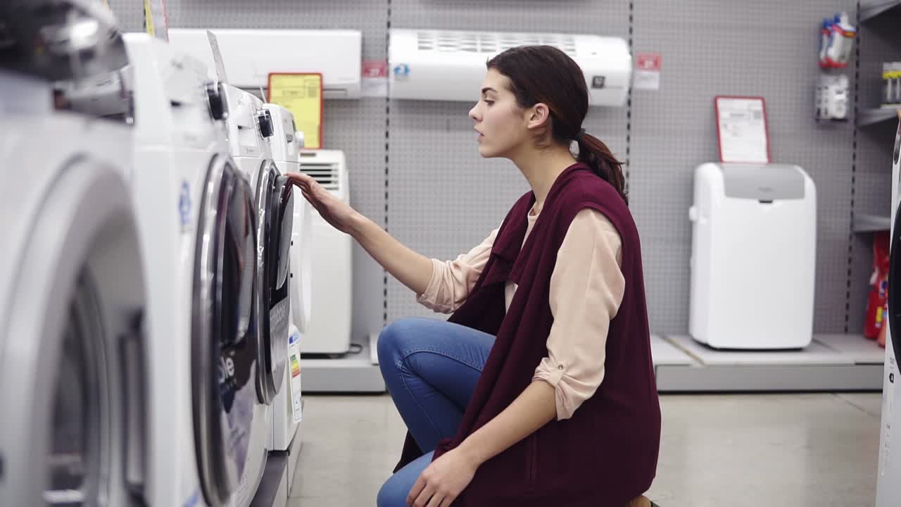 una joven y hermosa chica morena se baja para abrir una tapa de la lavadora en una tienda de electrodomésticos.