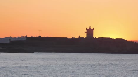 Tropischer-Carcavelos-meeresblick-Sonnenaufgang-über-Dem-Atlantik-Mit-Sonnenstrahlen,-Die-Sich-Durch-Das-Fort-Von-Saint-Julian-Bewegen,-Und-Kleinen-Wellen,-Die-Sich-Im-Vordergrund-Bei-Dem-Riesigen-Sonnenaufgang-Brechen