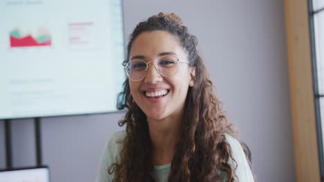 Portrait-of-smiling-biracial-businesswoman-wearing-glasses-in-meeting-room-with-screen-in-background