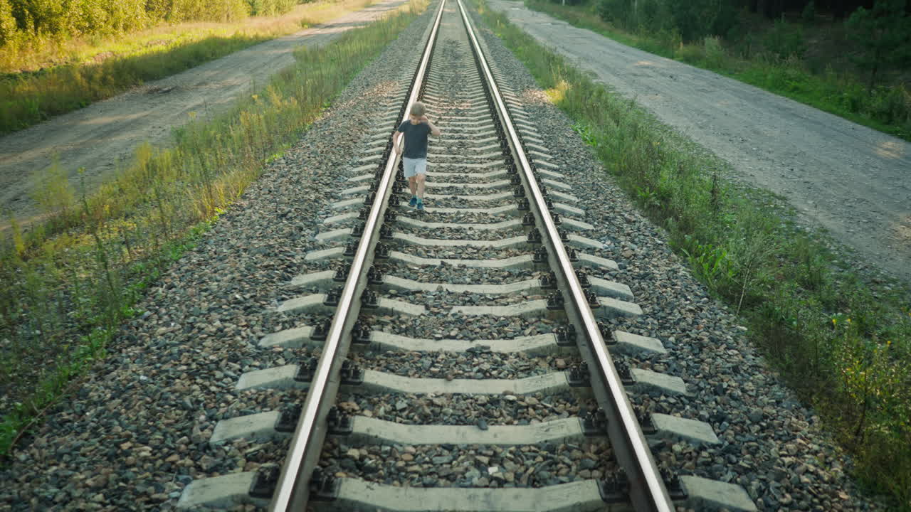 young boy in casual outfit walking alone on rail track in rural area, moving close to metal beam while scratching ear, surrounded by grassy terrain and dirt road on sunny da