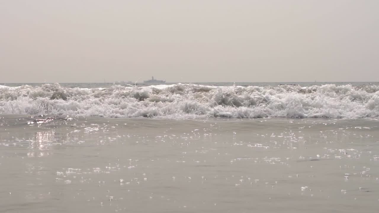 캘리포니아주 샌디에고의 모래사장에 파도가 슬로모션으로 부서지고 큰 배를 배경으로 - 와이드 샷