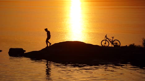 Male cyclist resting near river. Silhouette of young and active sportsman and his mountain bike standing near the river and looking far away at the sunset