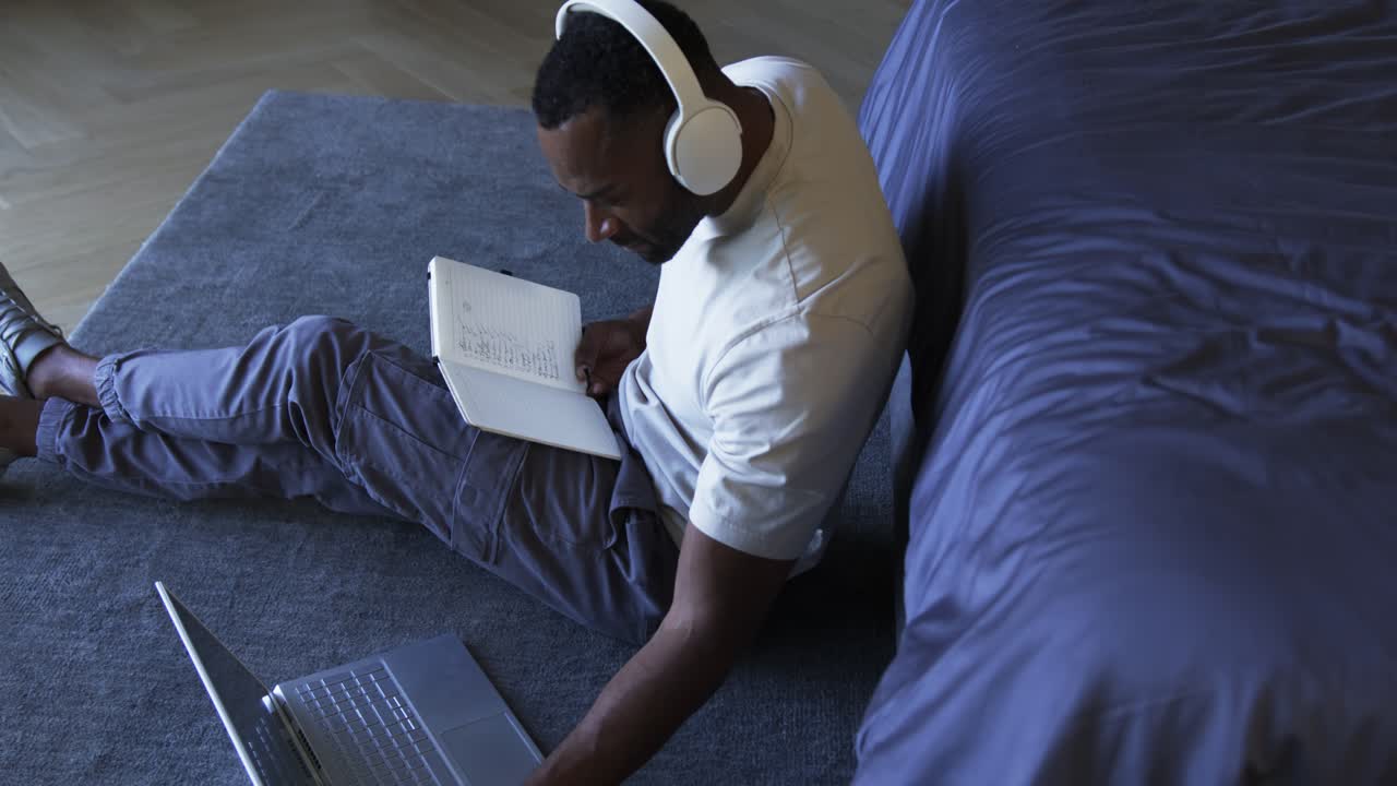 African American man consulting notebook, using laptop with headphones on rug by couch for work