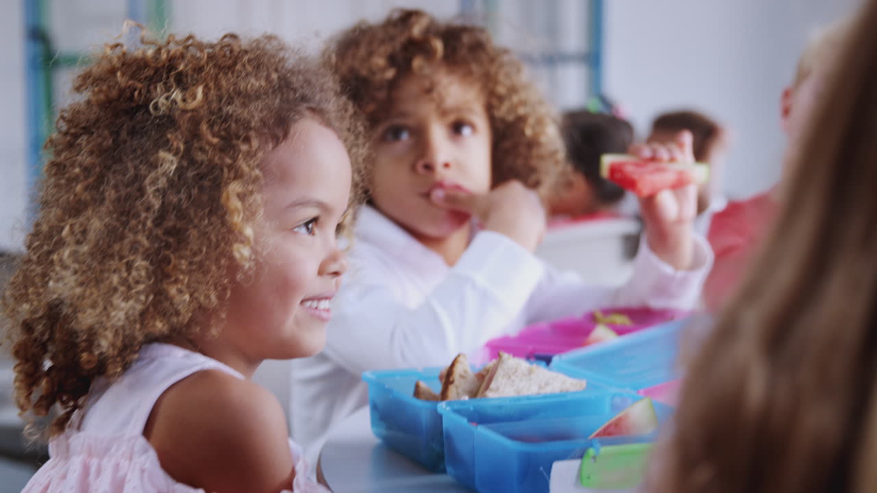 Bambina della scuola primaria a un tavolo con i compagni di classe che mangiano pranzi al sacco, messa a fuoco selettiva, primo piano
