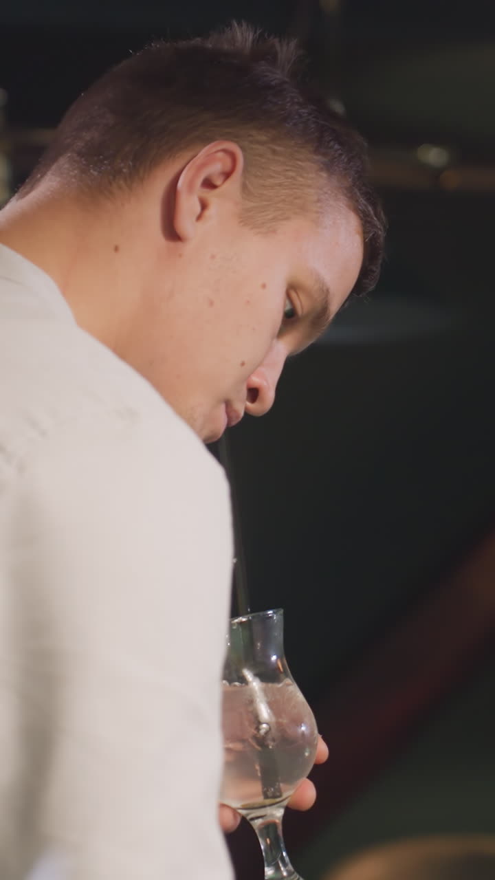 jugador de billar con camisa blanca bebiendo una bebida de limón con paja negra en una sala de billar débilmente iluminada. las luces superiores proyectan sombras dramáticas en la mesa de billar verde. expresión enfocada, ajuste de postura relajada