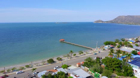 Drone-Shot-Forward-to-Playa-Juan-de-Bolanos-Town-Beach-Pier,-El-Muellecito-in-Summertime-and-Turquoise-Ocean-Waters,-Dominican-Republic