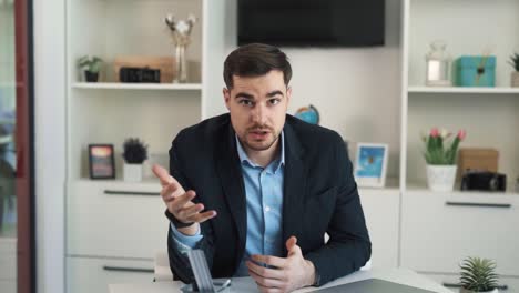 Portrait-of-an-angry-adult-man-in-business-attire-sitting-at-a-desk-with-a-laptop-in-a-beautiful,-bright-office-and-scolding-while-looking-into-the-camera