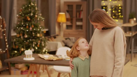 Young-Pretty-Woman-With-Toothy-Smile-Embracing-Her-Cute-Little-Daughter-While-Both-Looking-At-Camera-Against-Decorated-Firtree-At-Home