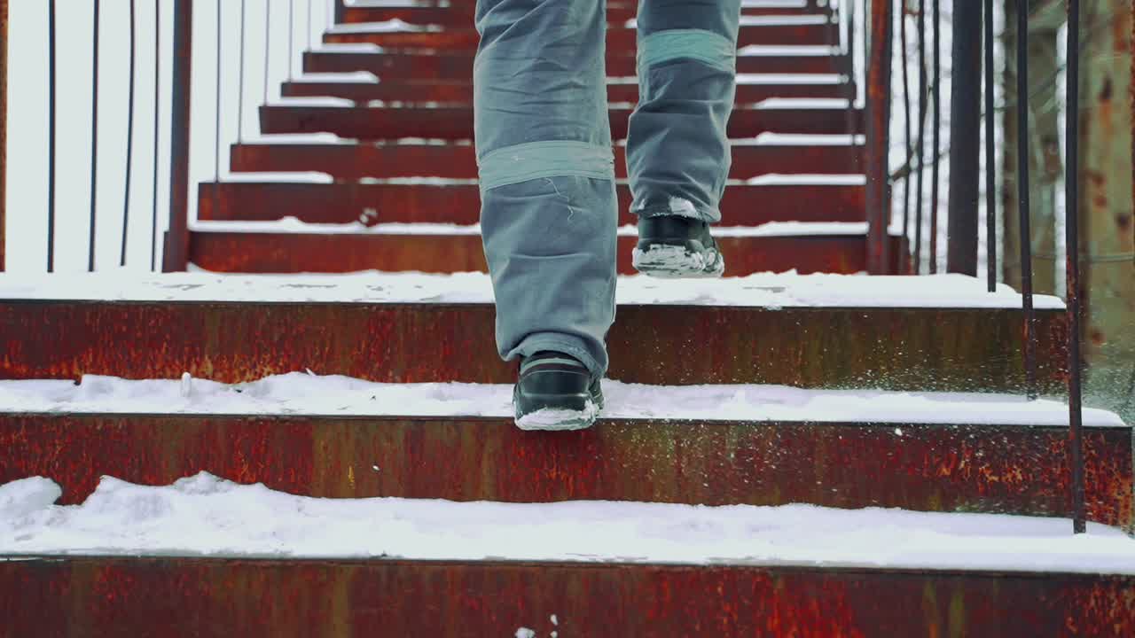 A worker in gray pants with a light horizontal stripe and black boots is climbing up steps in the street in the winter. Close-up of legs. View of back.