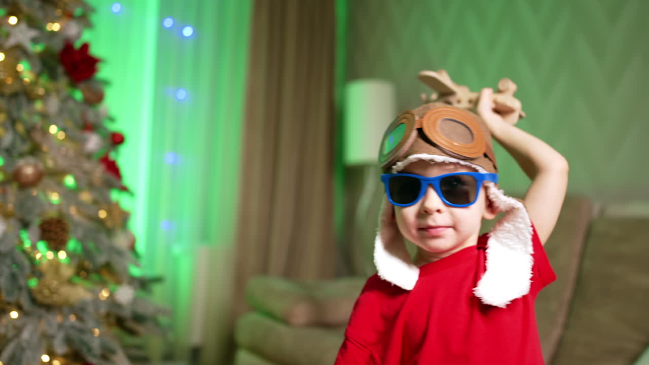 Cute toddler wearing pilot cap and sunglasses plays near the Christmas tree at home. Small kid plays with wooden airplane.
