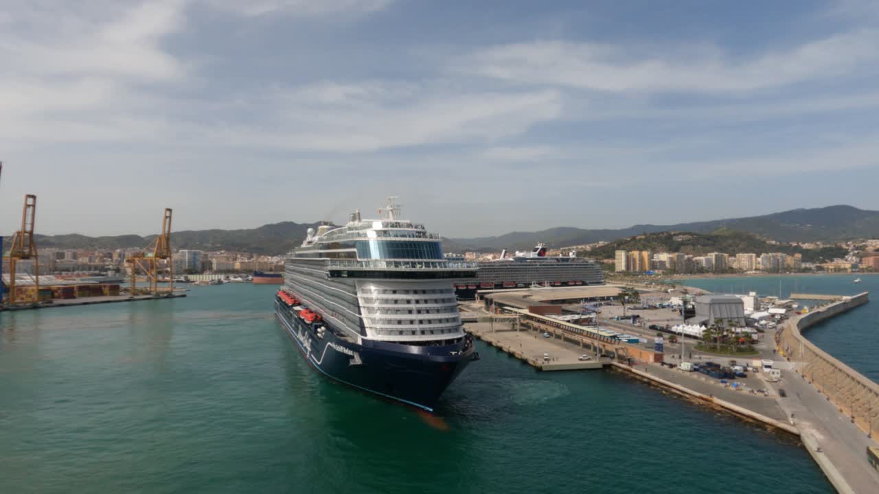 Stort krydstogtskib fortøjet i Malagas havn, Spanien. Luftperspektiv fra et andet fartøj afslører havneinfrastruktur, kraner og kystnært bybillede under blå himmel.