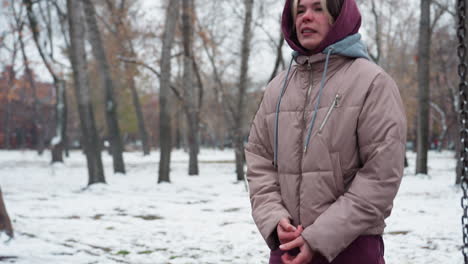 young woman in winter outfit feels cold, standing outside with snow on ground and trees in background, she appears chilly, bundled in warm winter gear