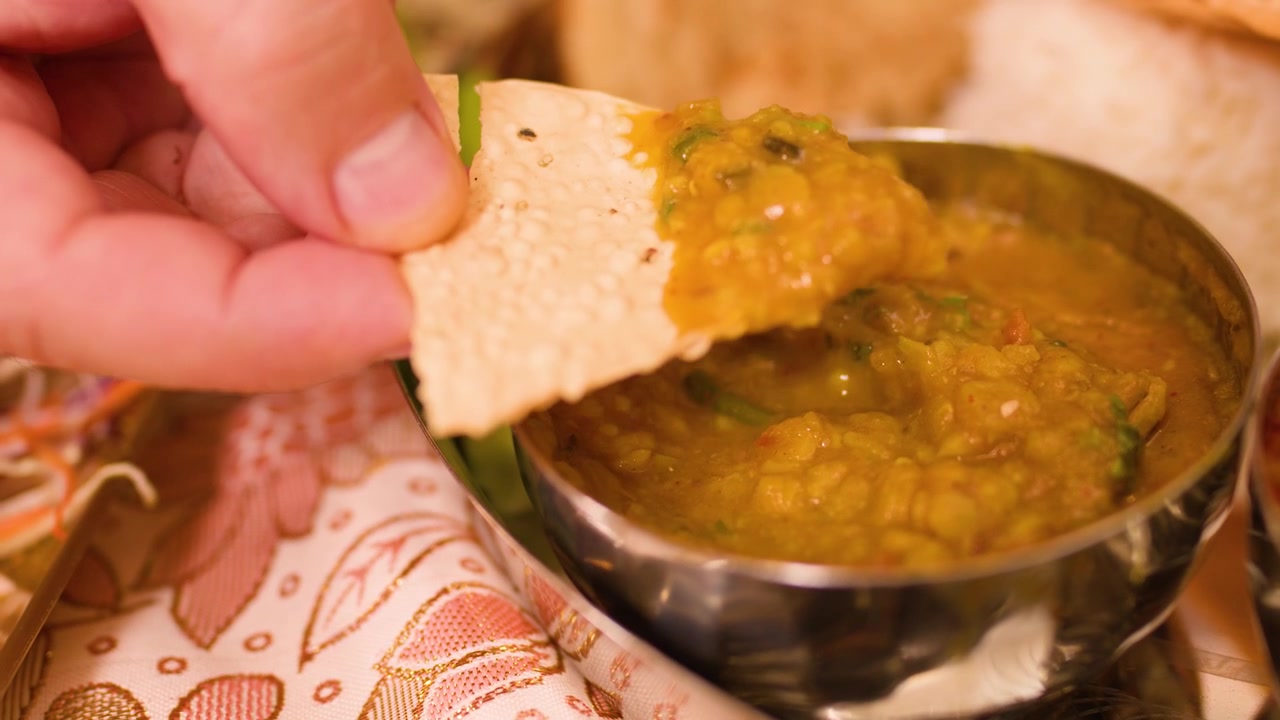 Närbild på hand som doppar krispig papadum i gul dalgryta i en metallskål.